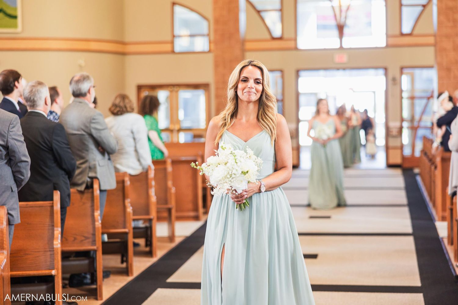 A woman in a seafoam green dress carries white flowers down a church aisle during a wedding ceremony.