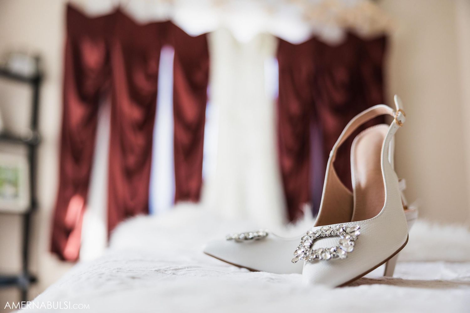 Elegant white bridal shoes with crystal embellishments rest on a bed with burgundy curtains in the background.