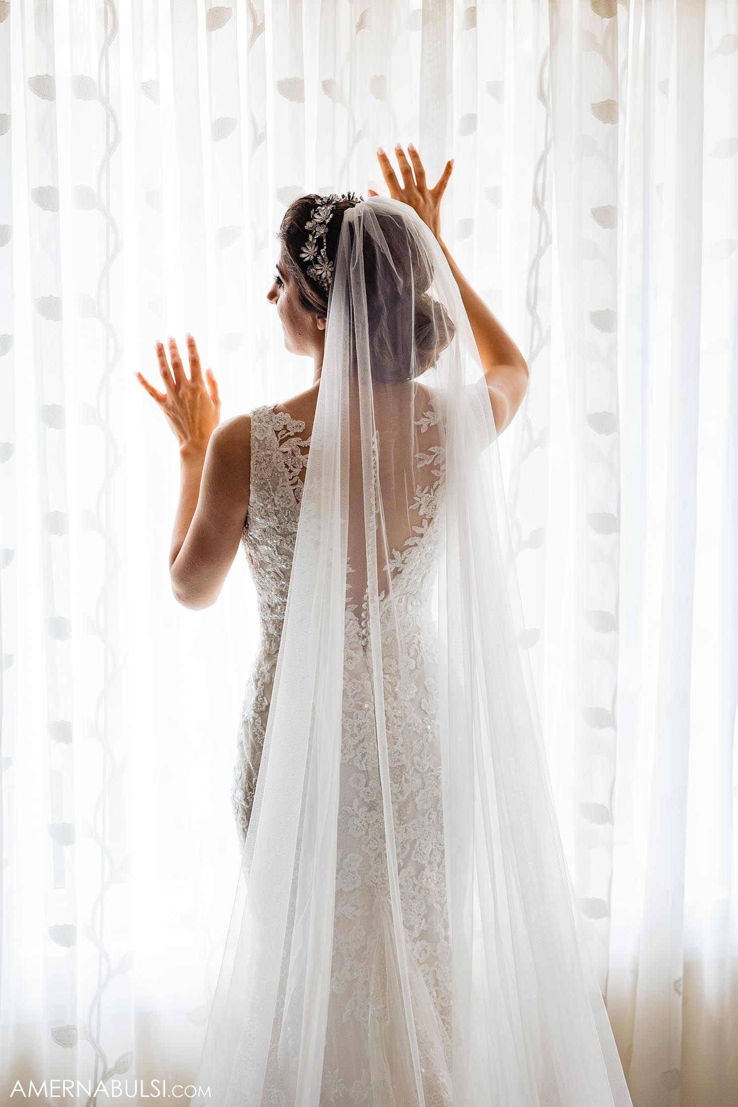 Elegant lace wedding dress against light window with flowing veil and delicate details.