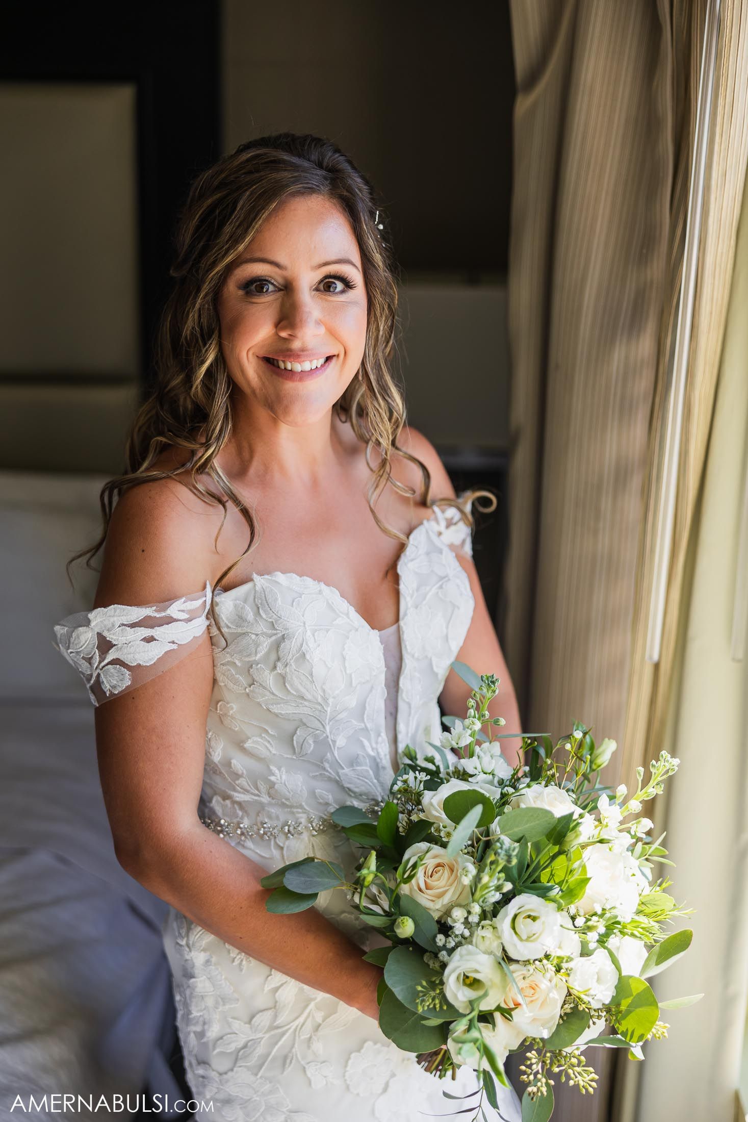 A wedding portrait showing a bride in lace dress holding a white and green bouquet by a window.