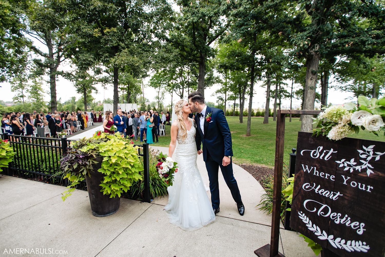 Wedding party walks along a tree-lined path in an outdoor garden setting.