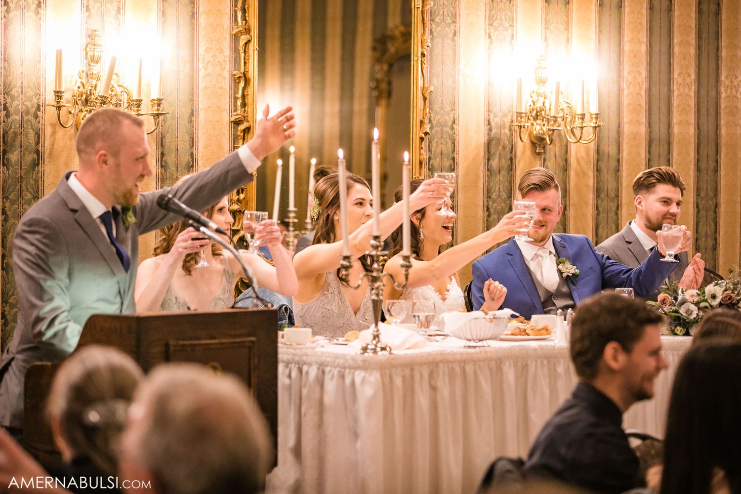 Wedding reception guests seated at elegant tables with chandeliers and warm lighting in a banquet hall.