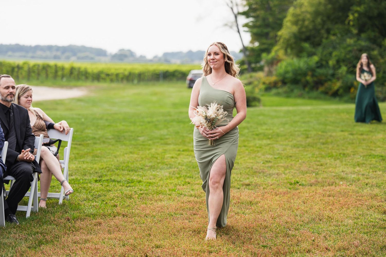 Bridesmaid walks down outdoor grassy aisle holding bouquet with wedding guests seated on chairs.