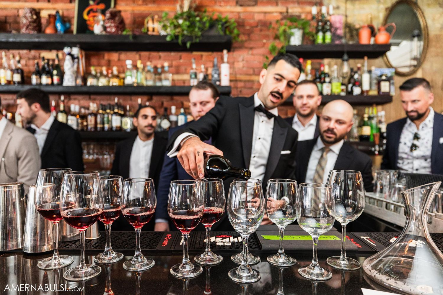 Collection of wine glasses lined up on a bar counter with people in formal attire.
