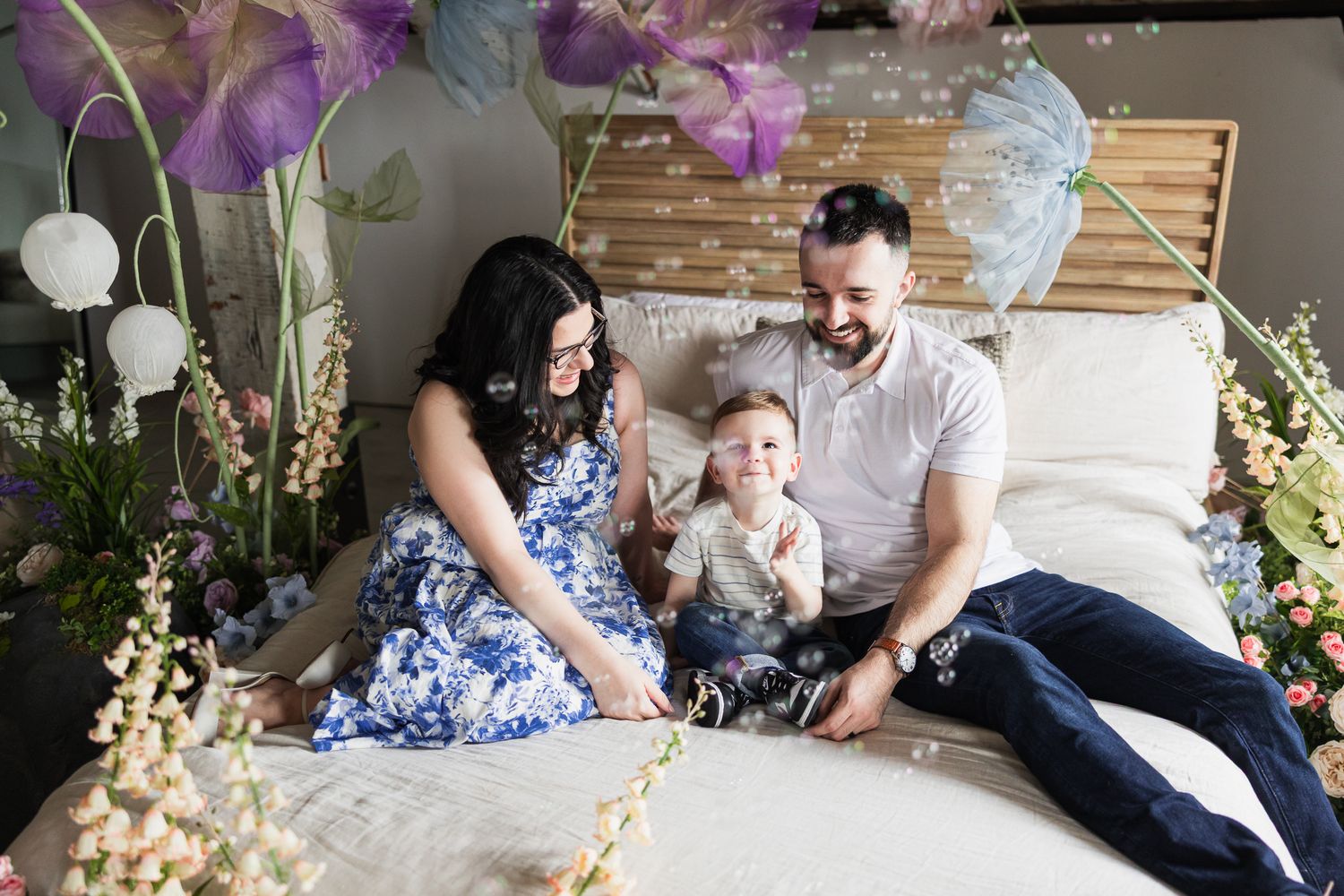 A family relaxes together on a white bed surrounded by colorful spring flowers and decorations.