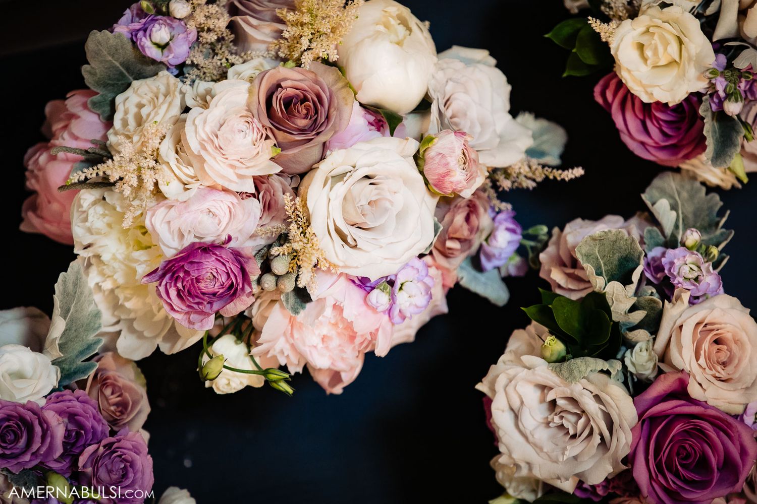 Elegant wedding bouquet featuring pink, cream and purple roses arranged against dark background.