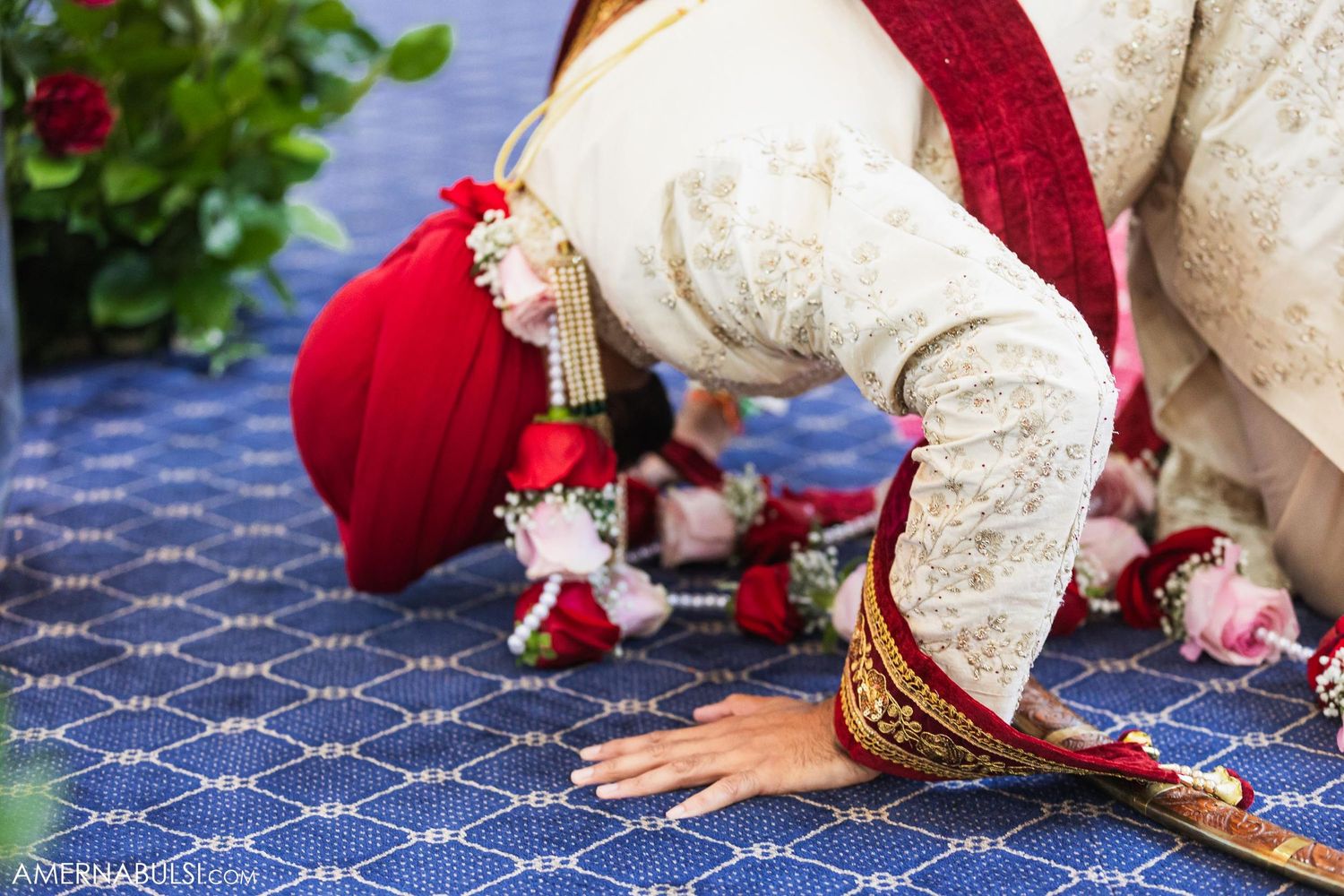 Traditional Sikh ceremony participants kneeling on blue patterned carpet wearing red and cream colored attire.
