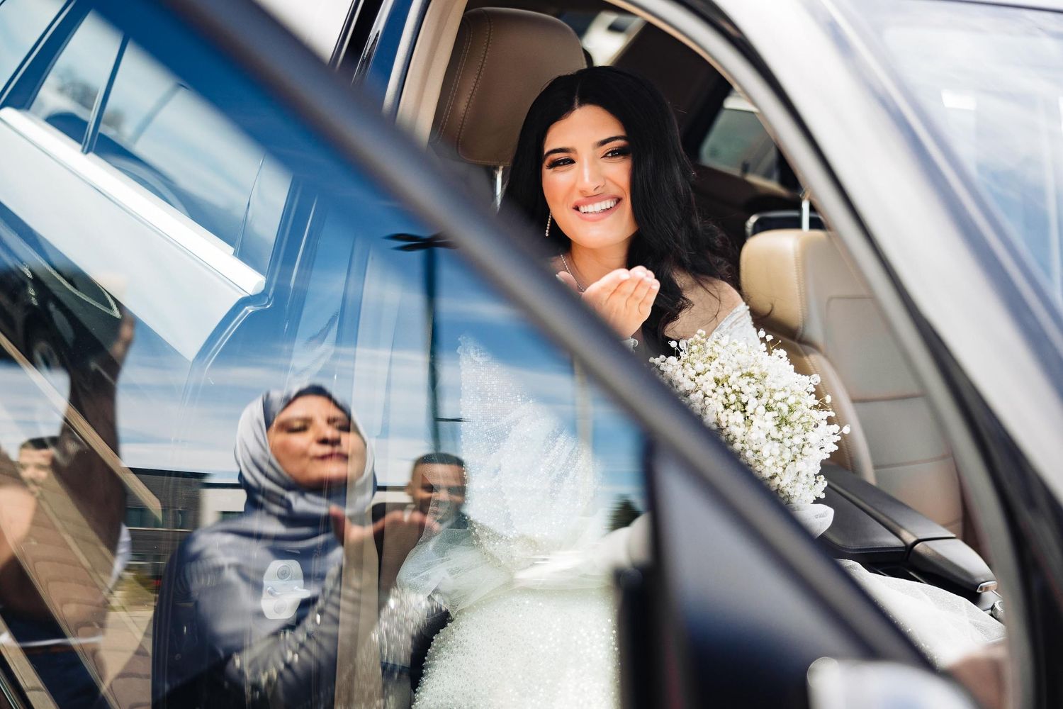 Joyful bride sits in car with beautiful bouquet while being photographed through window.