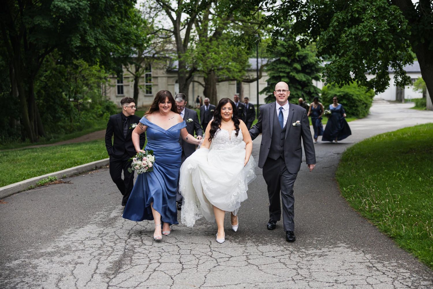 Wedding party walking together on garden path surrounded by green trees.