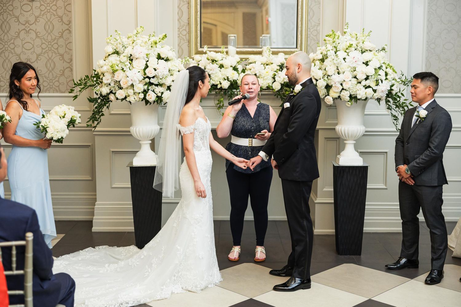 Wedding ceremony in progress with elegant white floral arrangements and checkerboard flooring.