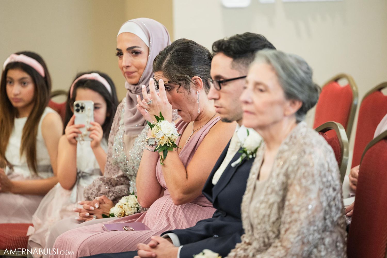 Wedding guests watch ceremony intently from red chairs in an indoor venue.