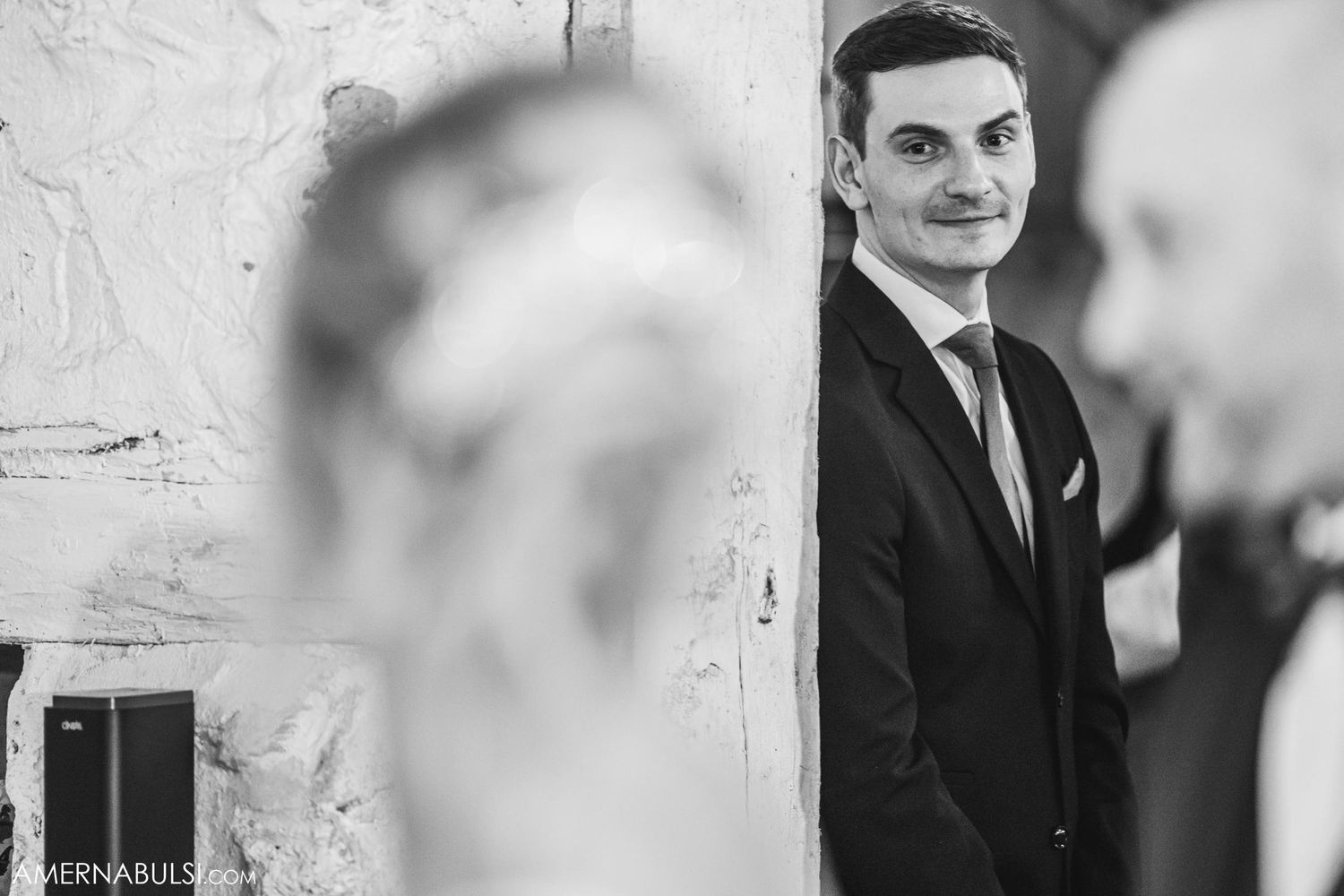 A well-dressed man stands in profile against a white textured wall at a formal event.