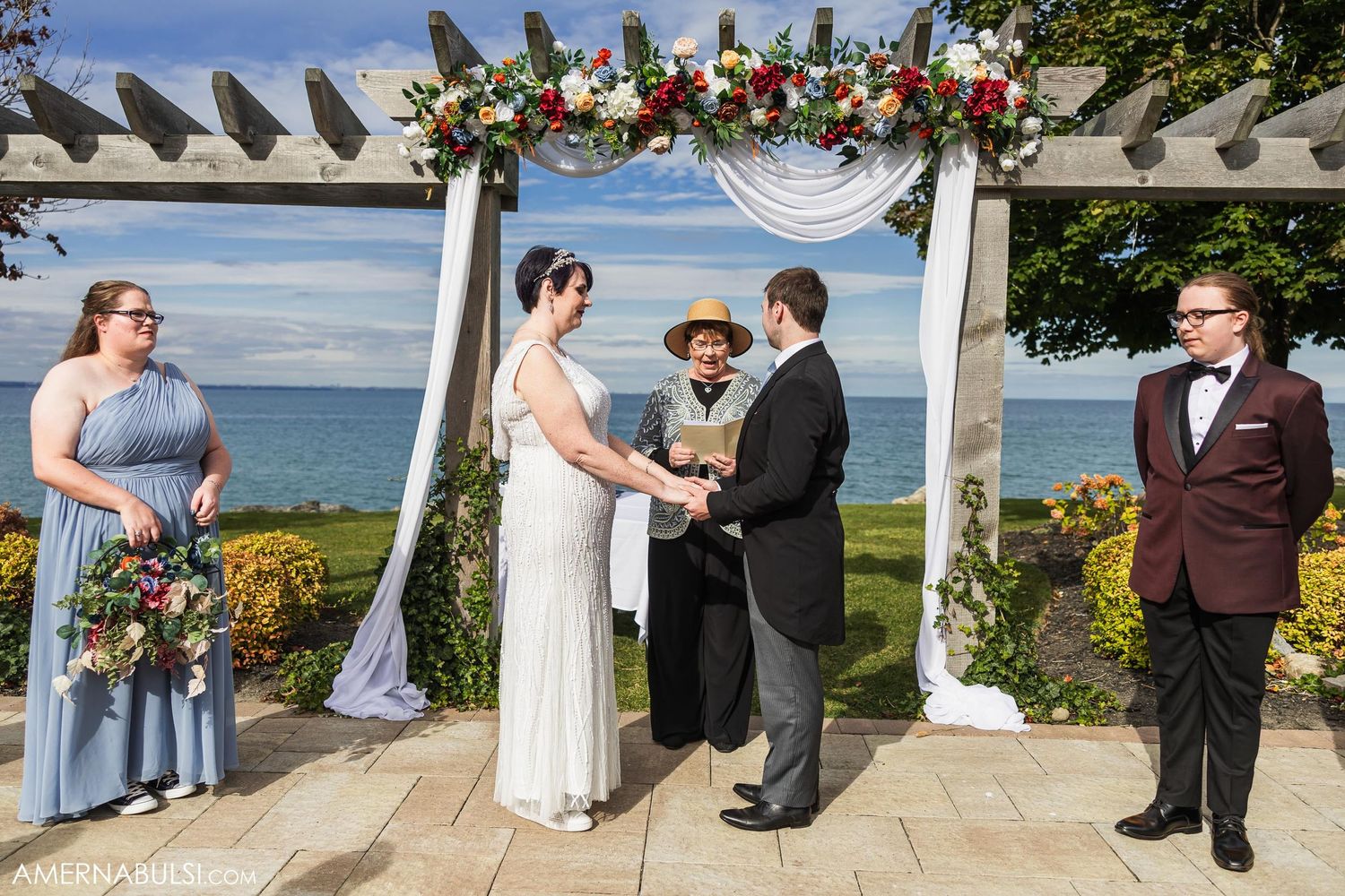 Outdoor waterfront wedding ceremony under a floral-decorated pergola with bridal party.