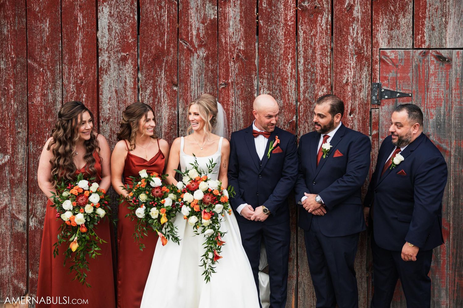 Wedding party poses together in front of rustic red barn wall with beautiful floral bouquets.