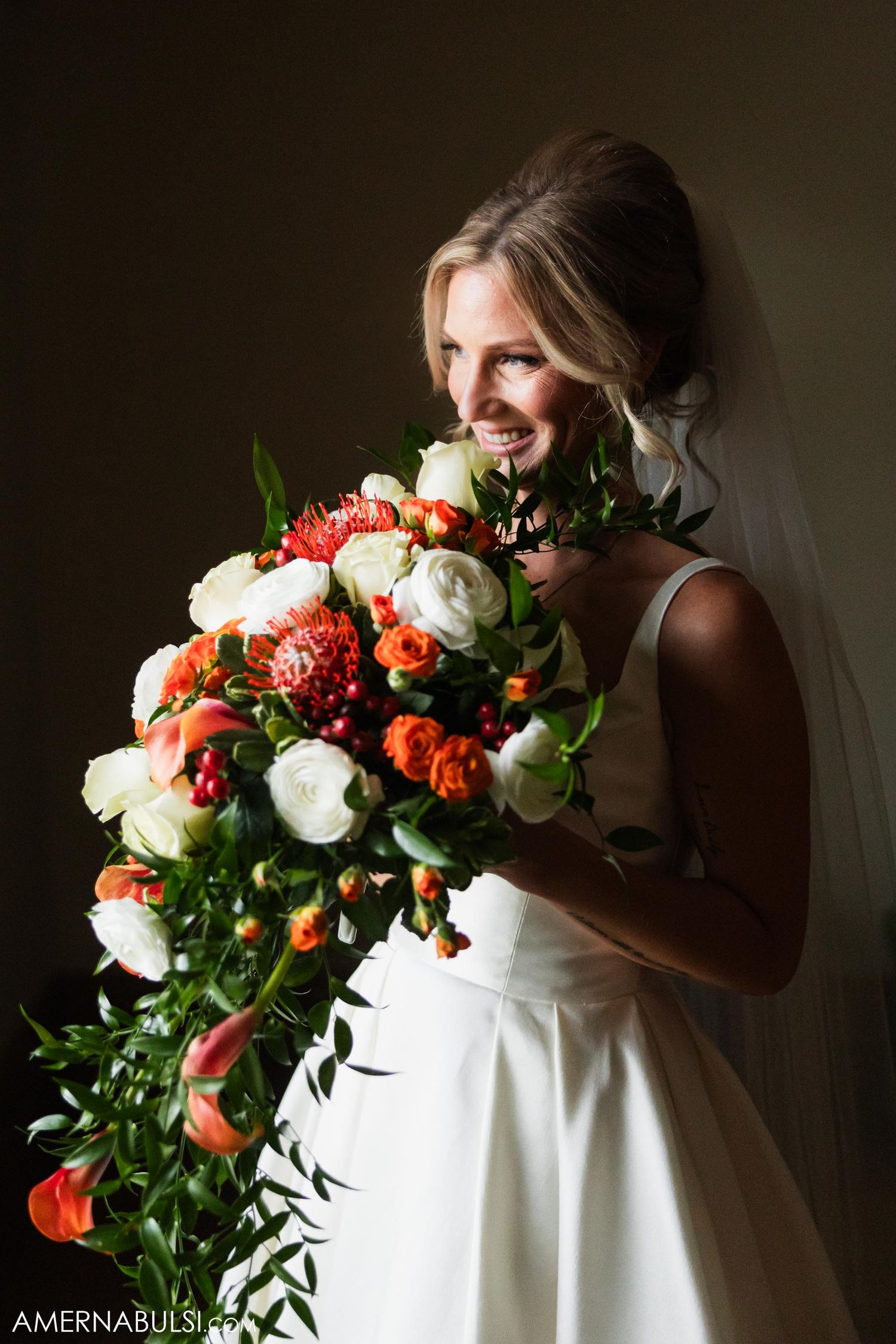 A bride holds a vibrant bouquet with coral and white flowers against a dark background.