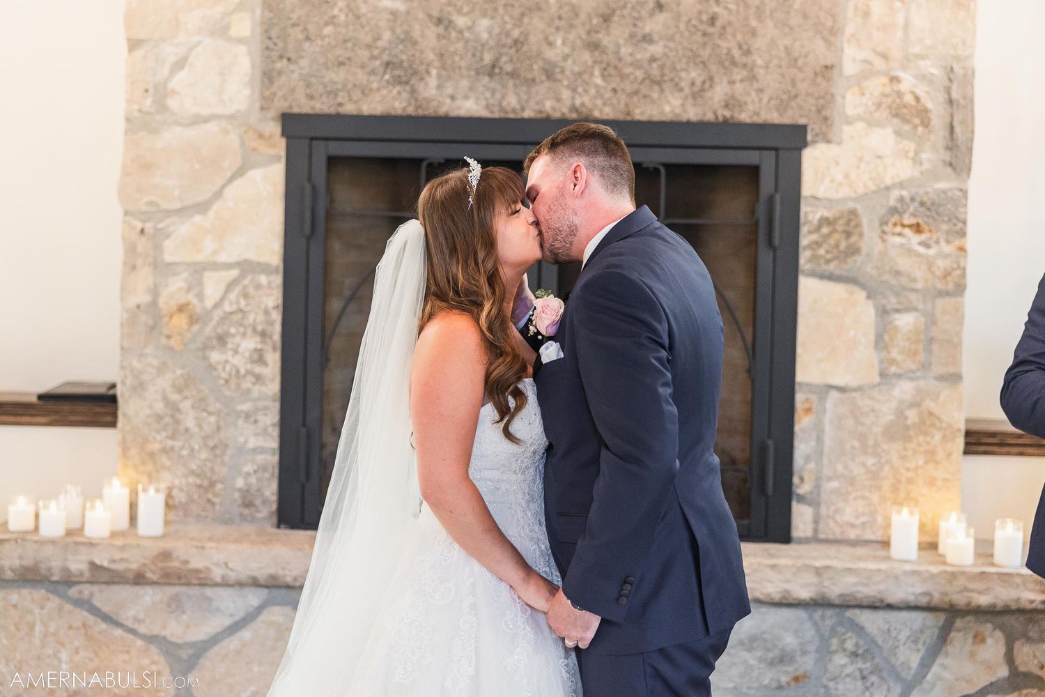 Romantic first kiss as newlyweds in front of stone fireplace with candles.