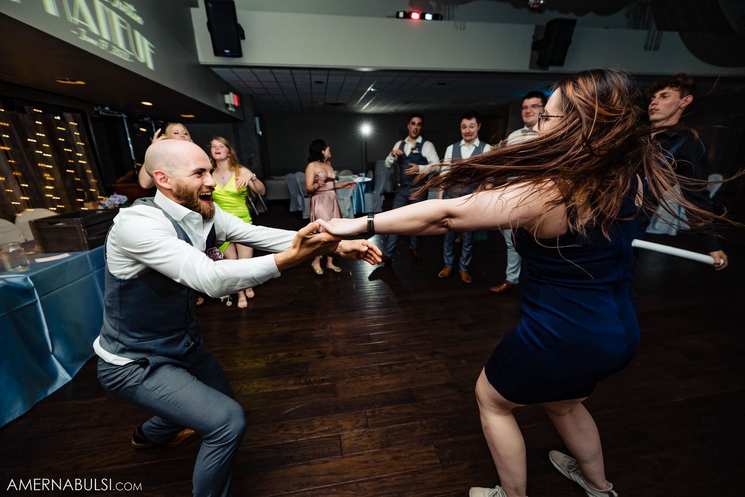 Enthusiastic wedding guests dancing and reaching across dance floor during lively reception moment.