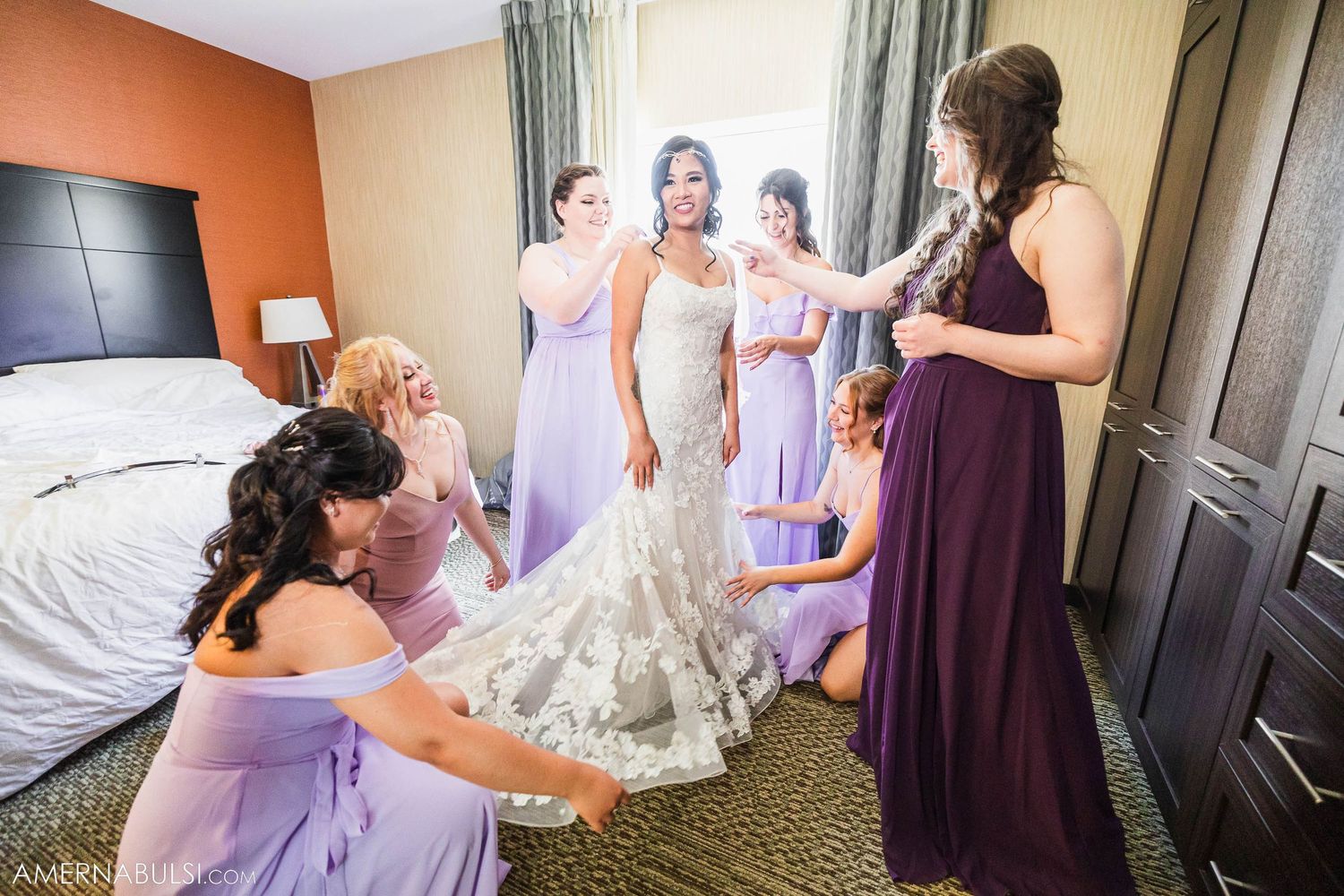 Wedding party helps adjust bride's dress in hotel room.
