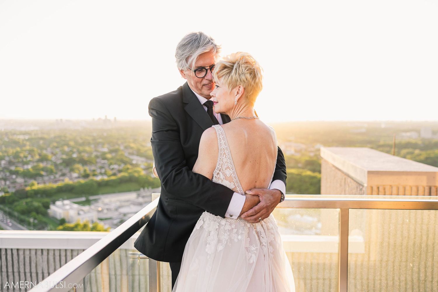 A couple in wedding attire shares a tender moment on a balcony with a cityscape backdrop.
