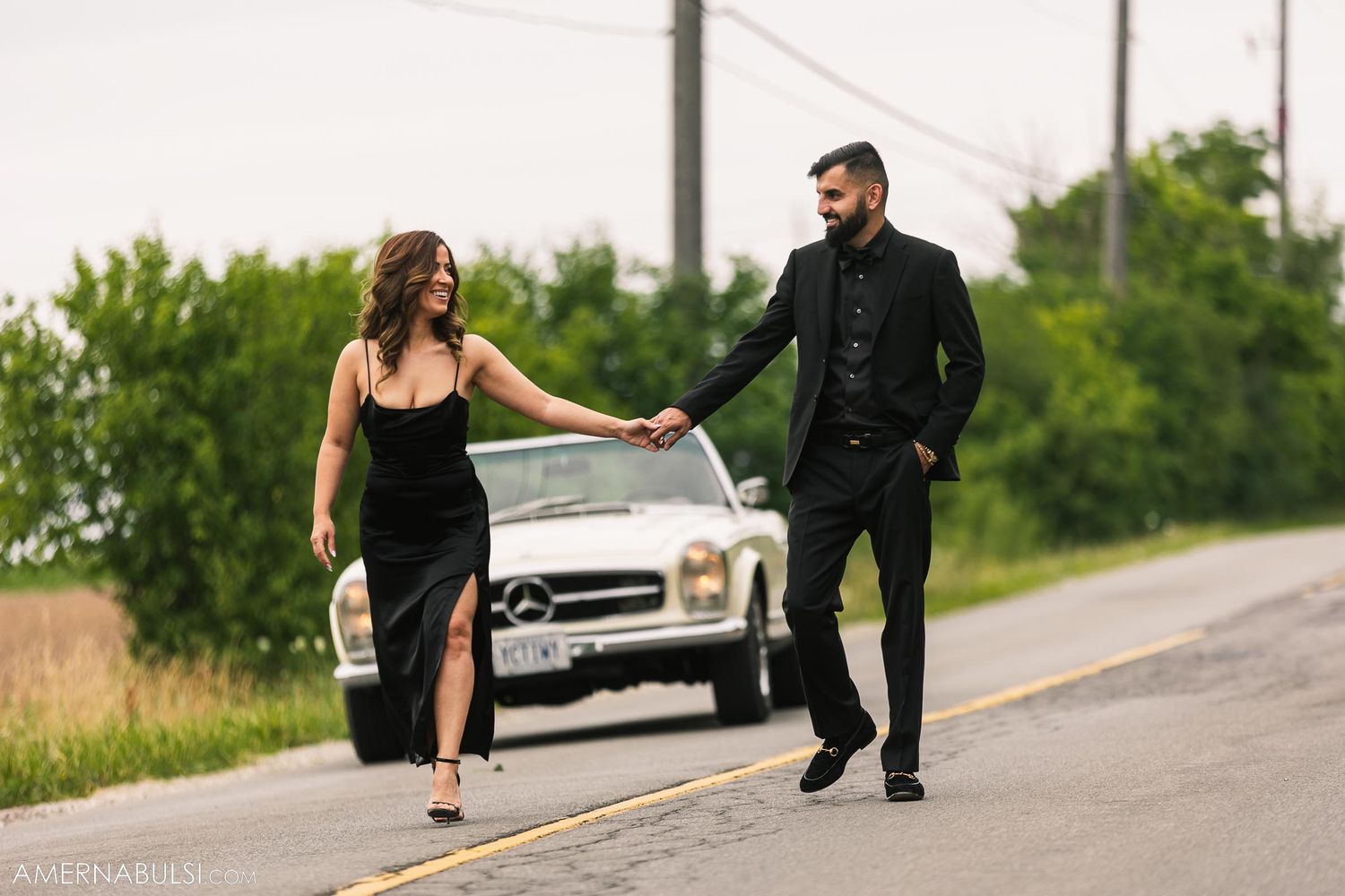 Stylish couple walking together on a rural road with a parked car in the background.