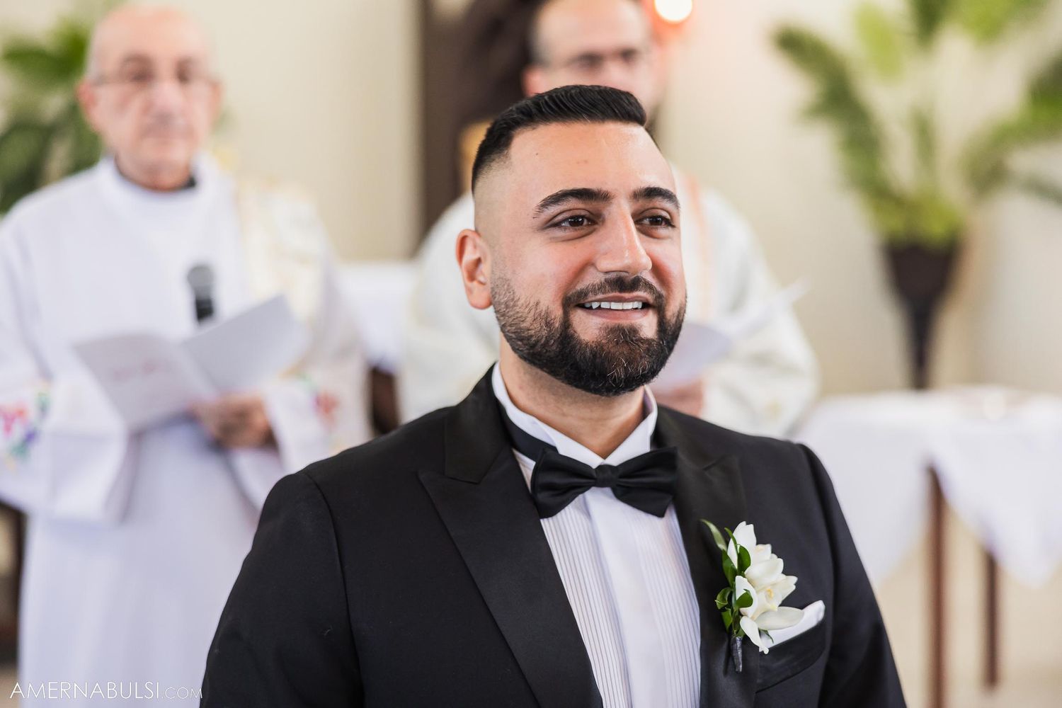 A groom wearing a black tuxedo and bowtie smiles during the wedding ceremony.