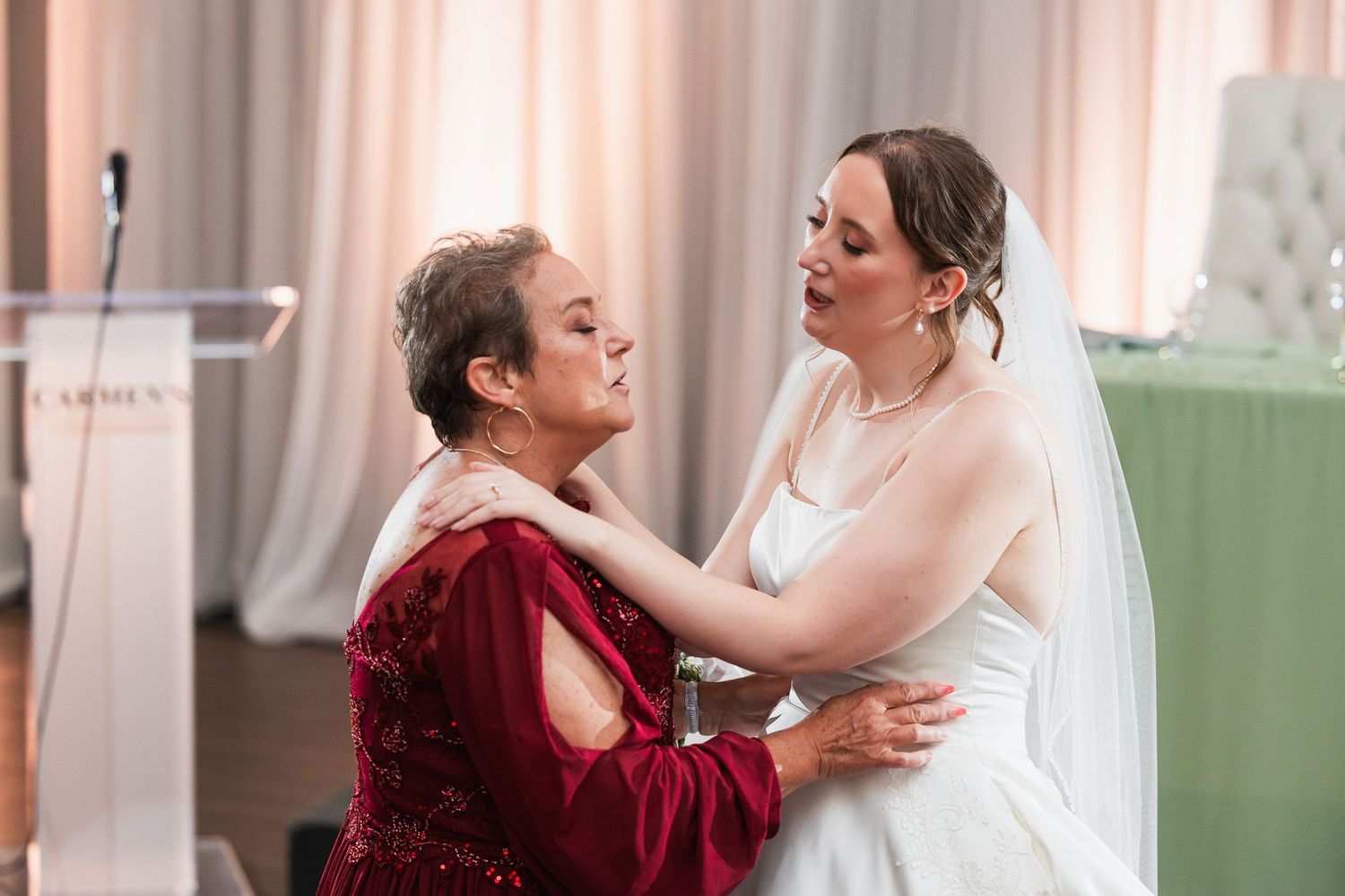 Bride in white gown and veil shares emotional moment with woman in red dress at wedding reception.