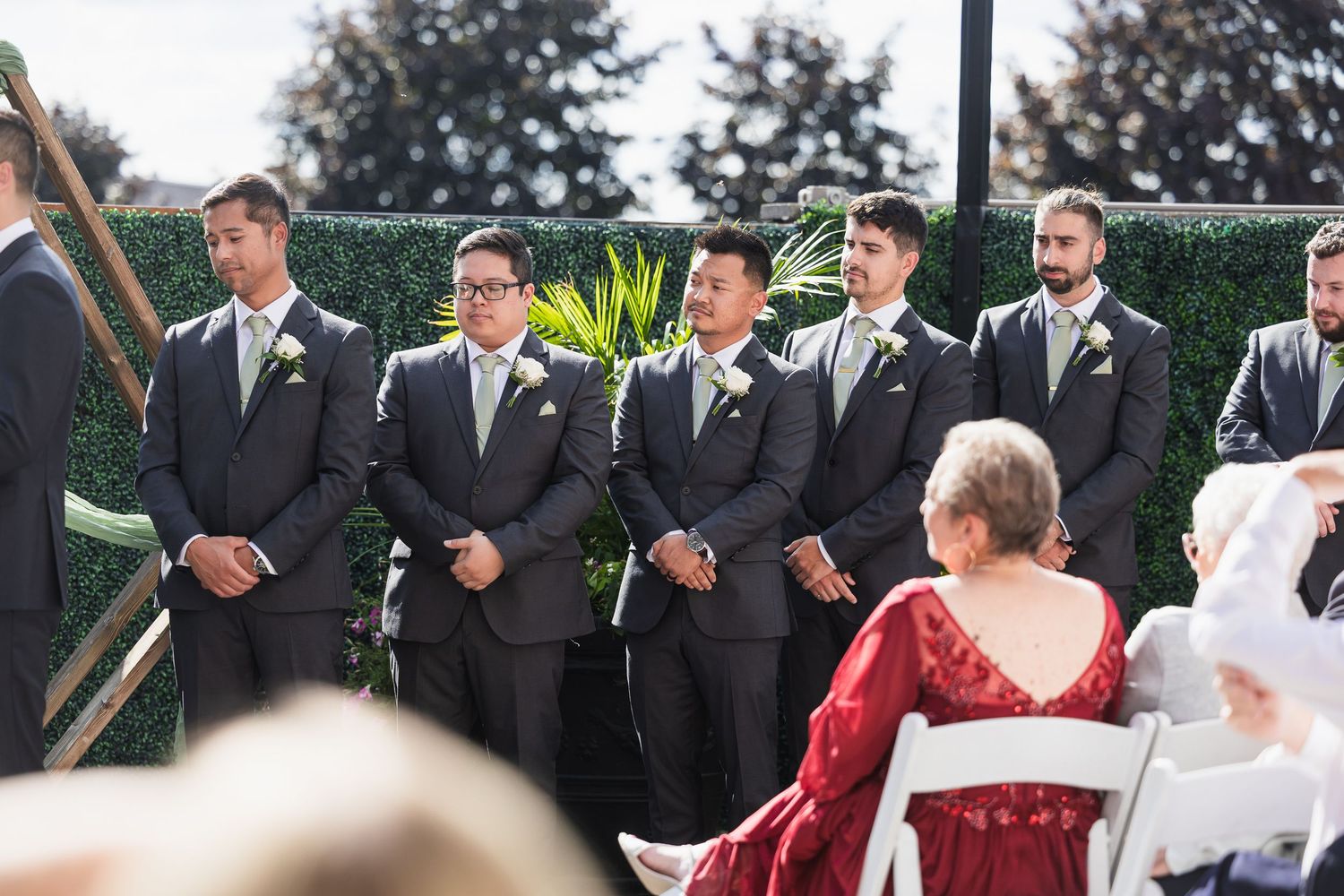 Groomsmen in gray suits with boutonnieres stand at outdoor wedding ceremony beside lush green hedge backdrop.