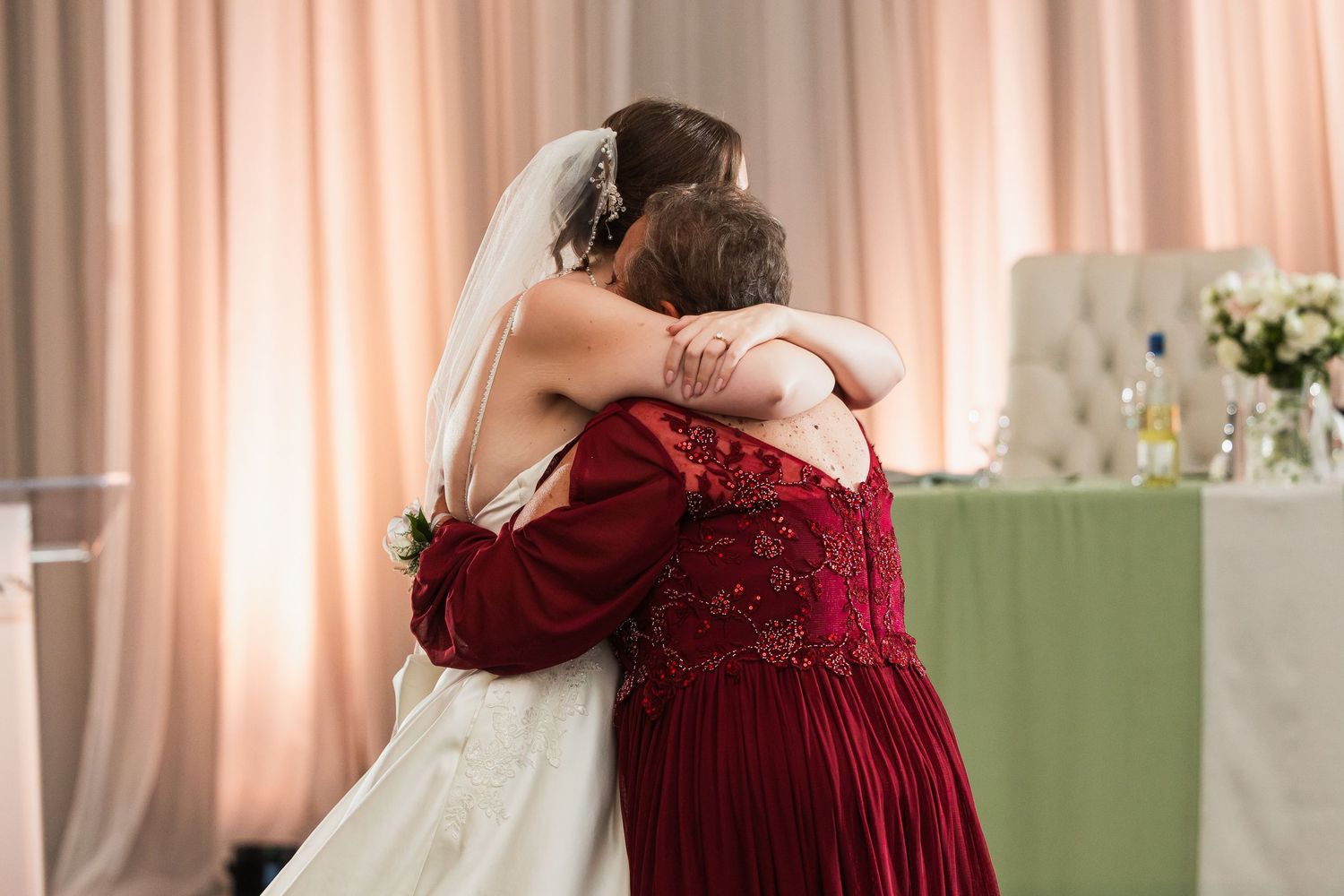 Bride and mother sharing an emotional hug in wedding reception venue with pink draping backdrop.