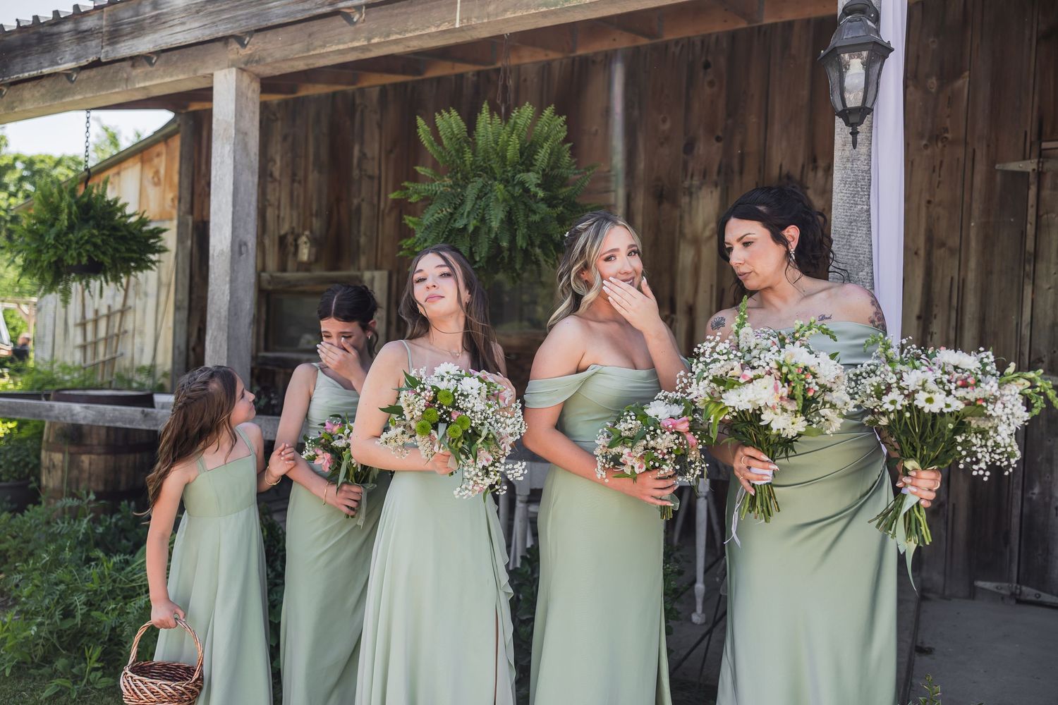 Bridesmaids in sage green gowns holding white floral bouquets outside a rustic barn wedding venue.