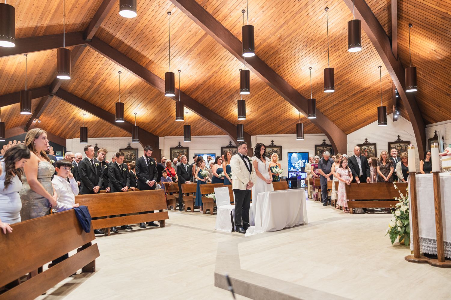 Wedding ceremony in a wooden-ceiling Catholic church with guests seated in pews as couple stands at altar.