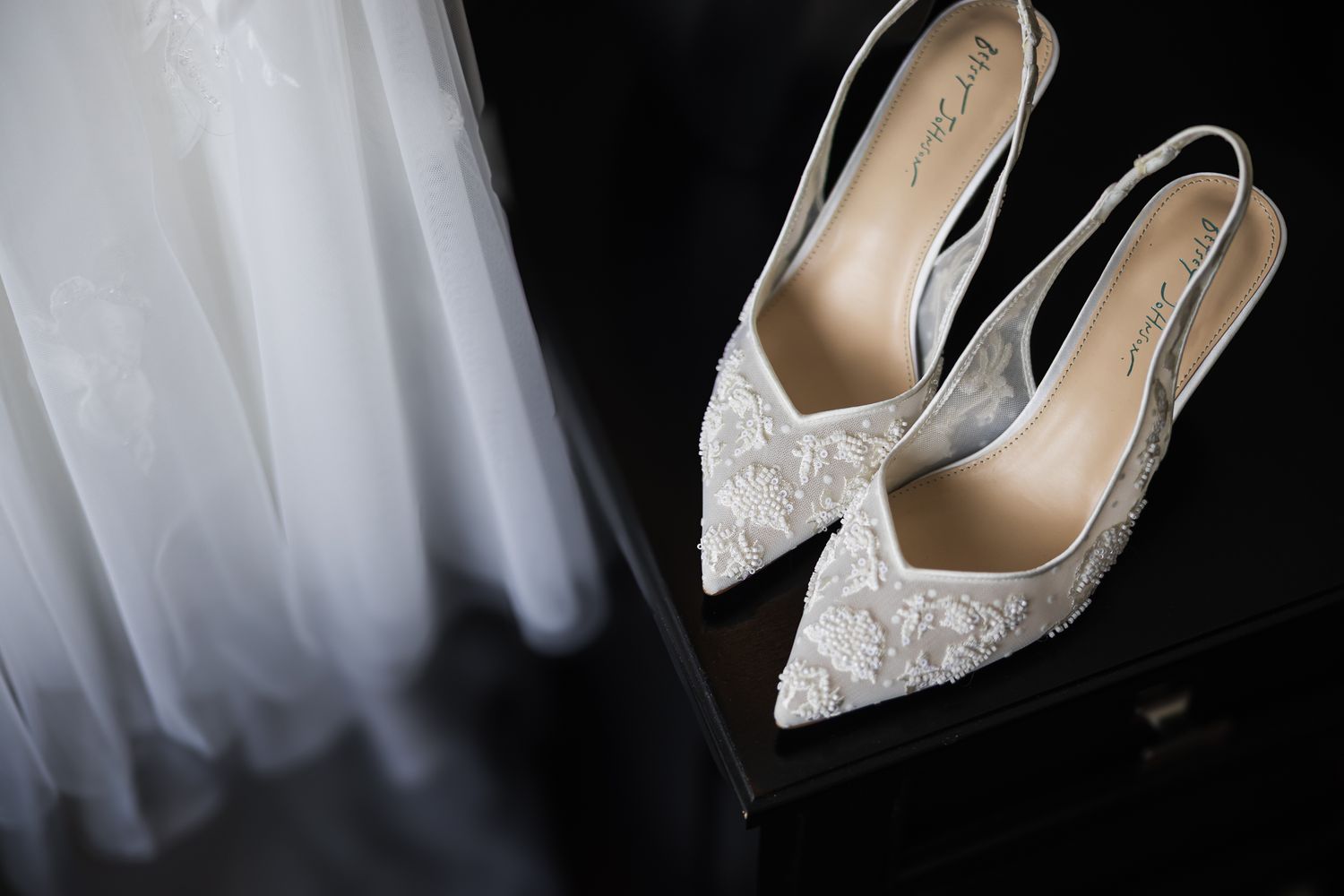 Elegant white lace pointed-toe slingback wedding heels displayed on a dark dresser beside a bridal gown.