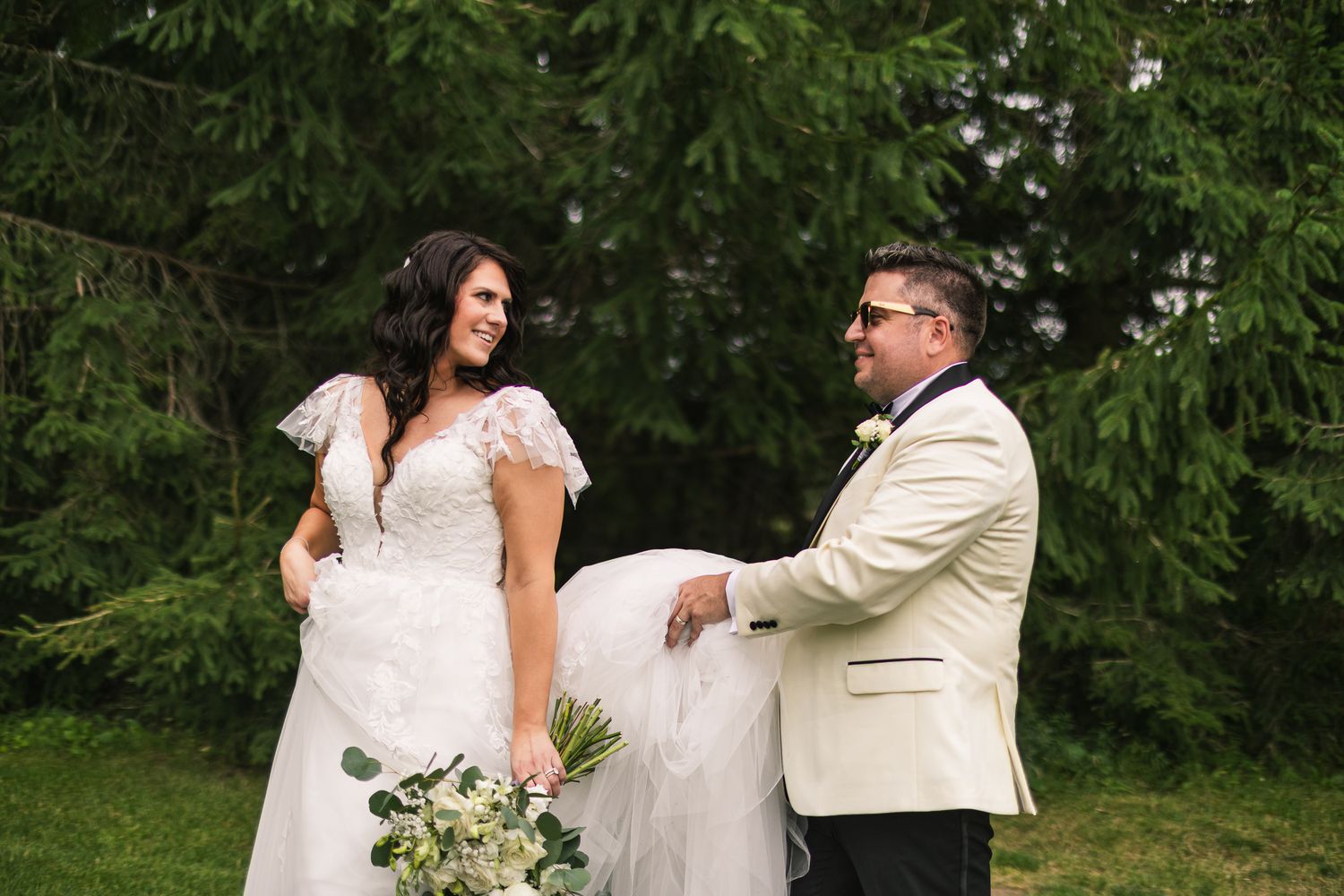 Groom in cream jacket helps bride with her white lace wedding gown train outdoors near evergreen trees.