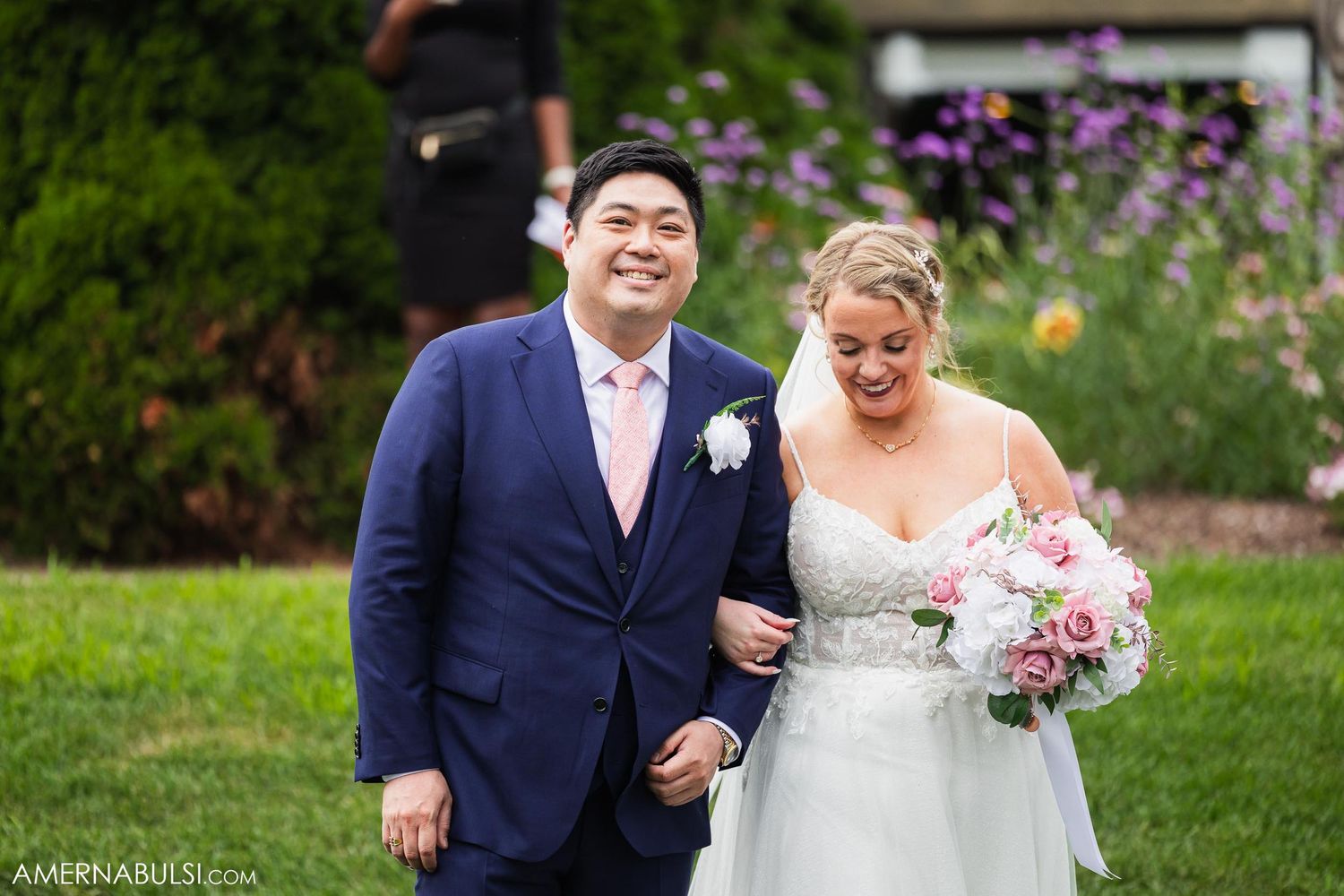Wedding couple in navy suit and white dress holding bouquet of pink flowers in garden setting.