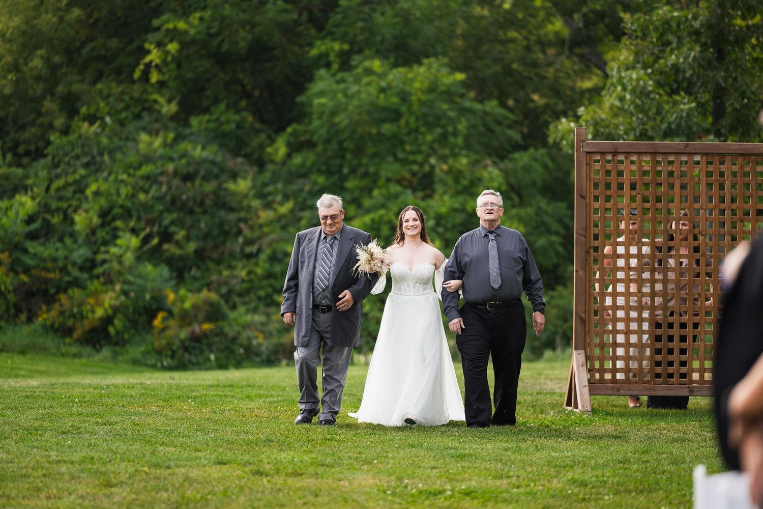 Bride walks down outdoor aisle with two escorts past wooden lattice structure.