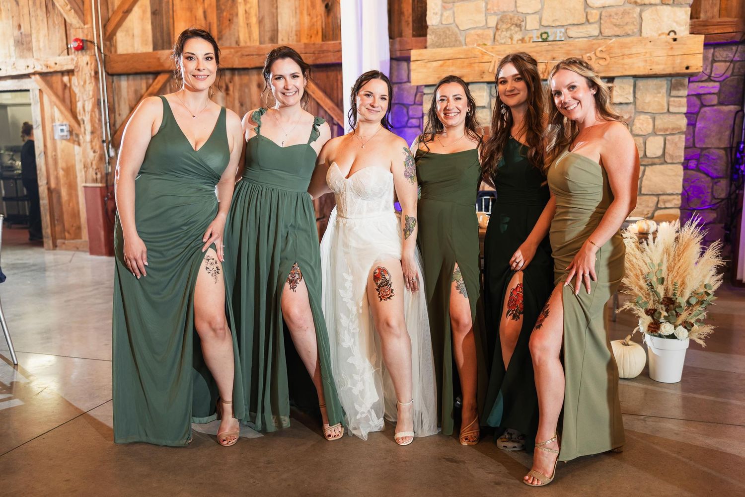Bridesmaids wearing matching sage green dresses pose with the bride in a rustic barn venue.