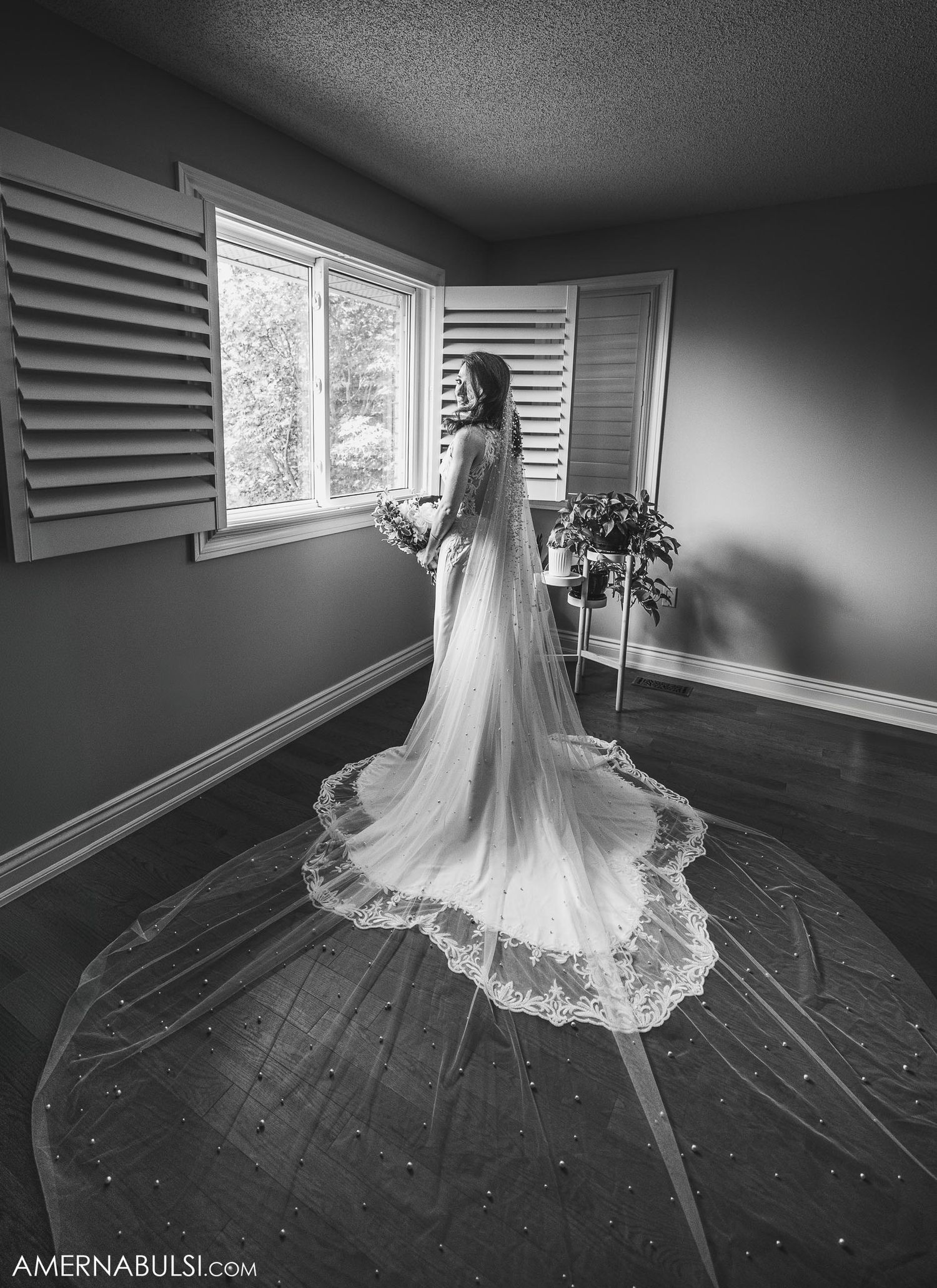 Dramatic black and white photo of a wedding dress with long lace train by a window with shutters.
