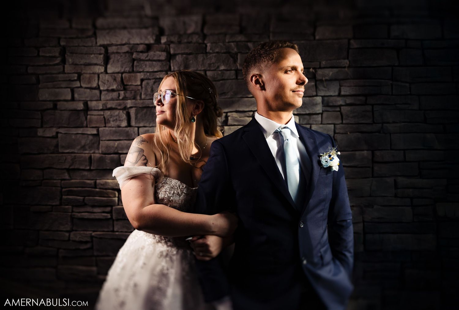 A couple shares a romantic moment in front of a dark textured brick wall showing elegant wedding attire.
