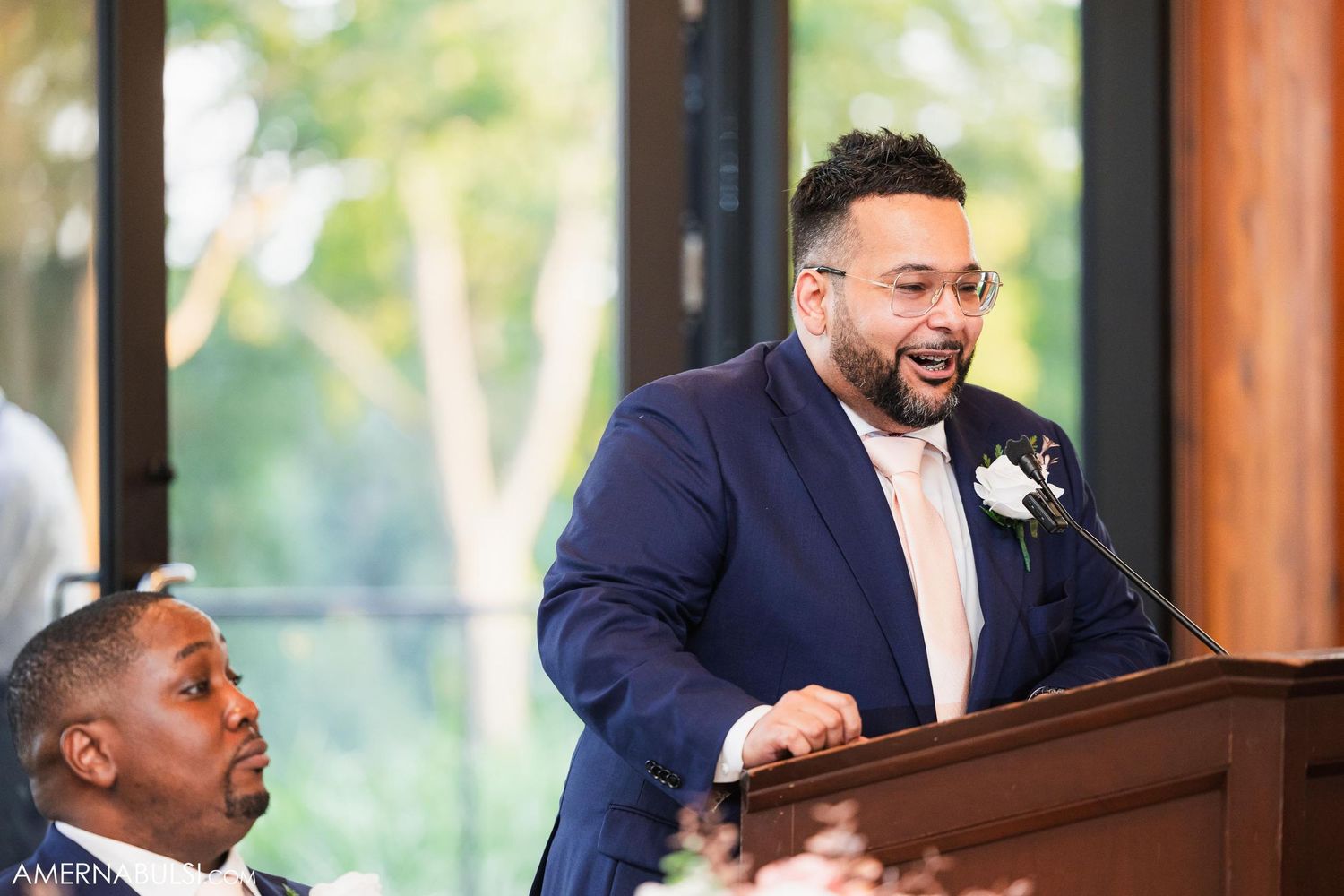 A speaker delivers a heartfelt speech at a wedding reception podium while wearing a navy suit.