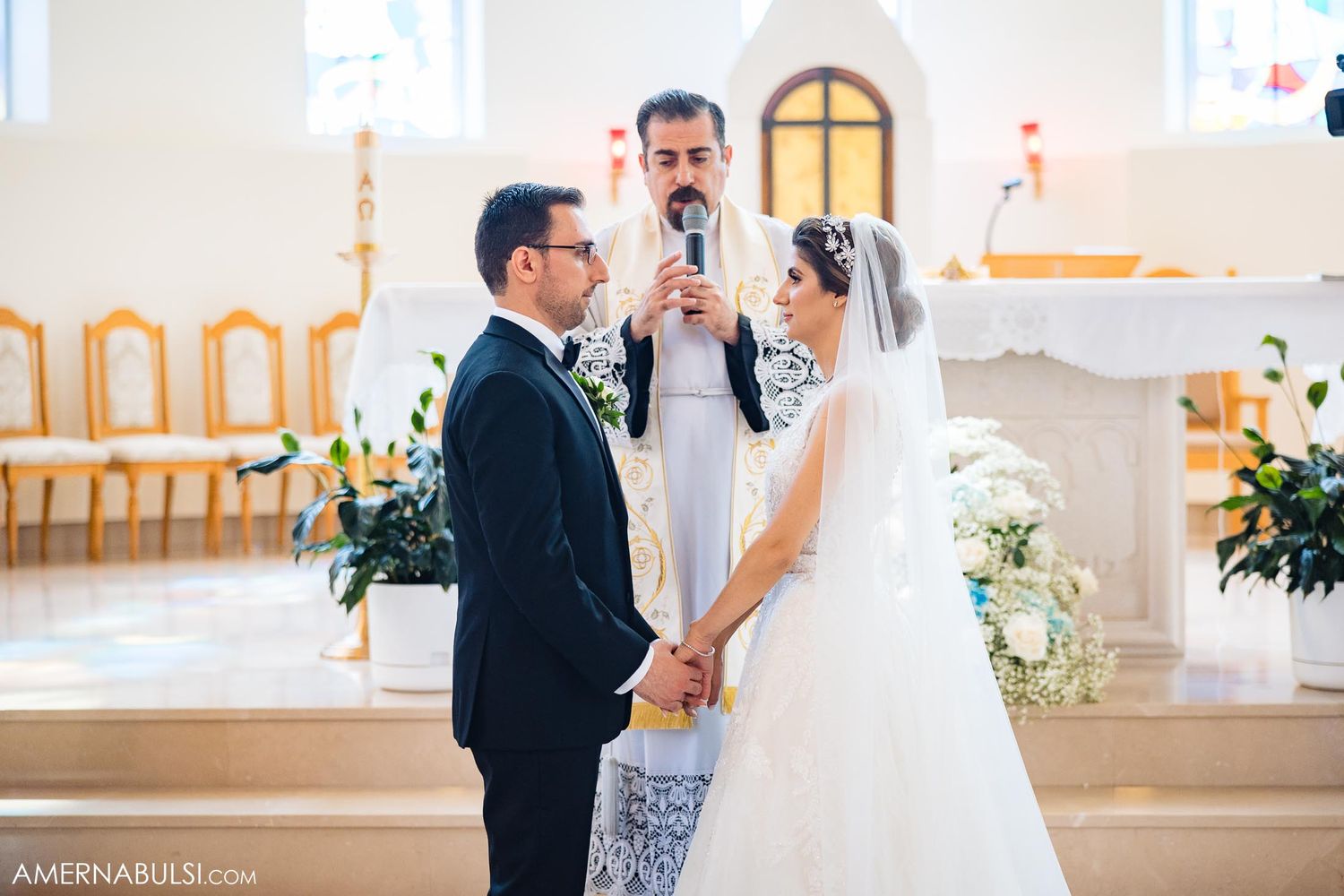 Wedding ceremony in progress with priest officiating as couple holds hands at the altar.