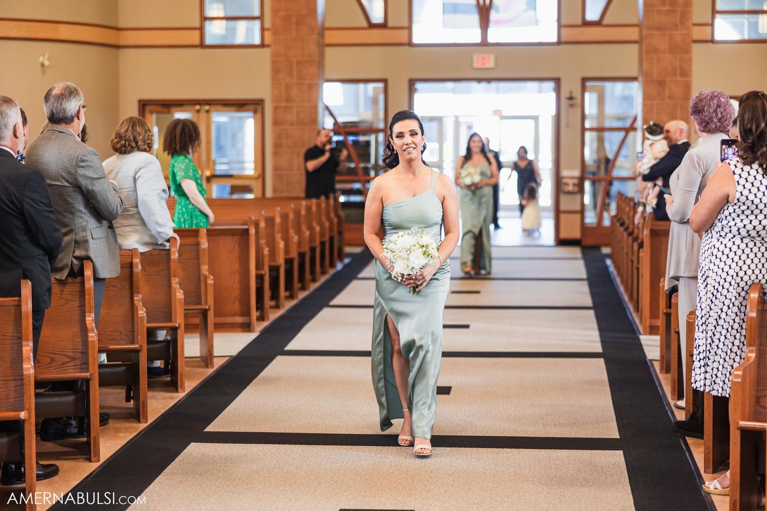 A bridesmaid in a light blue dress walks down the church aisle holding a bouquet during a wedding ceremony.