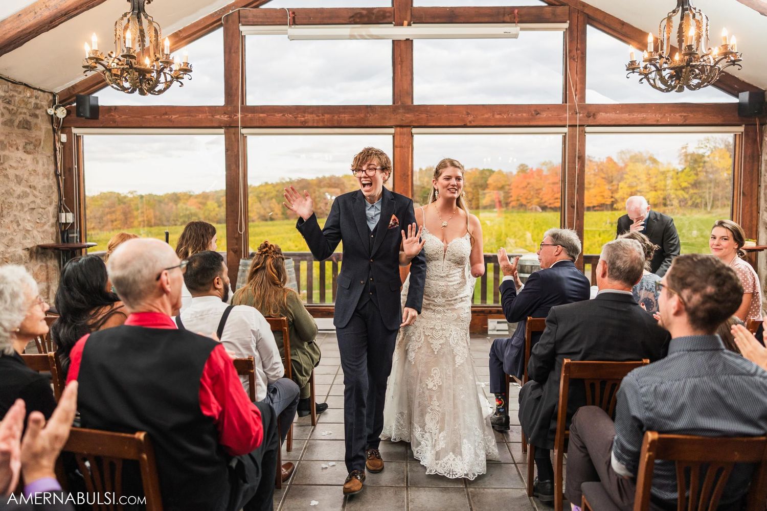 Wedding couple stands triumphantly before seated guests in rustic barn venue with fall landscape views.