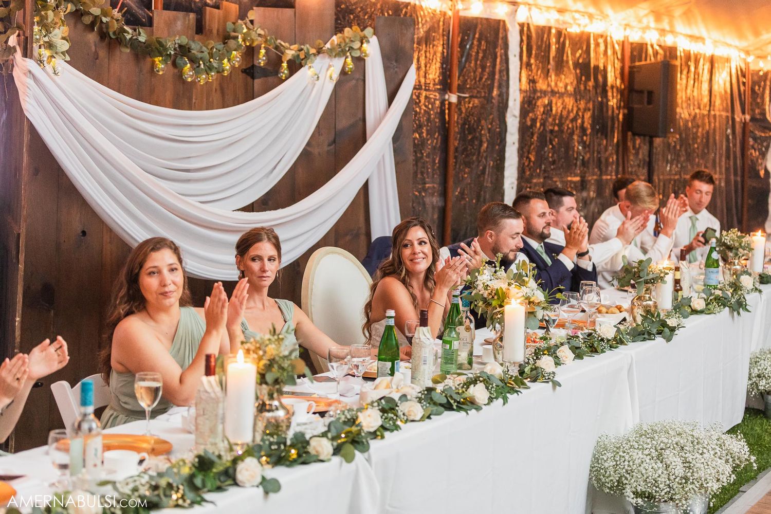 A wedding reception head table decorated with white draping and floral garlands glows in warm lighting.