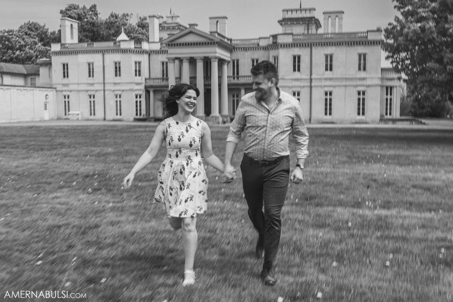 Black and white photo of couple strolling playfully in front of historic mansion.