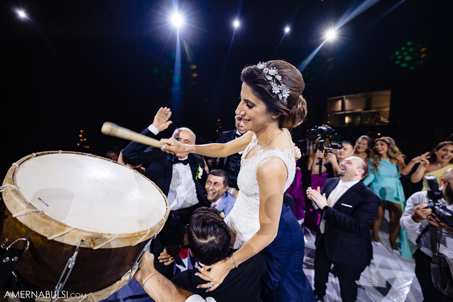 A bride in white dress plays a large drum during wedding reception festivities.