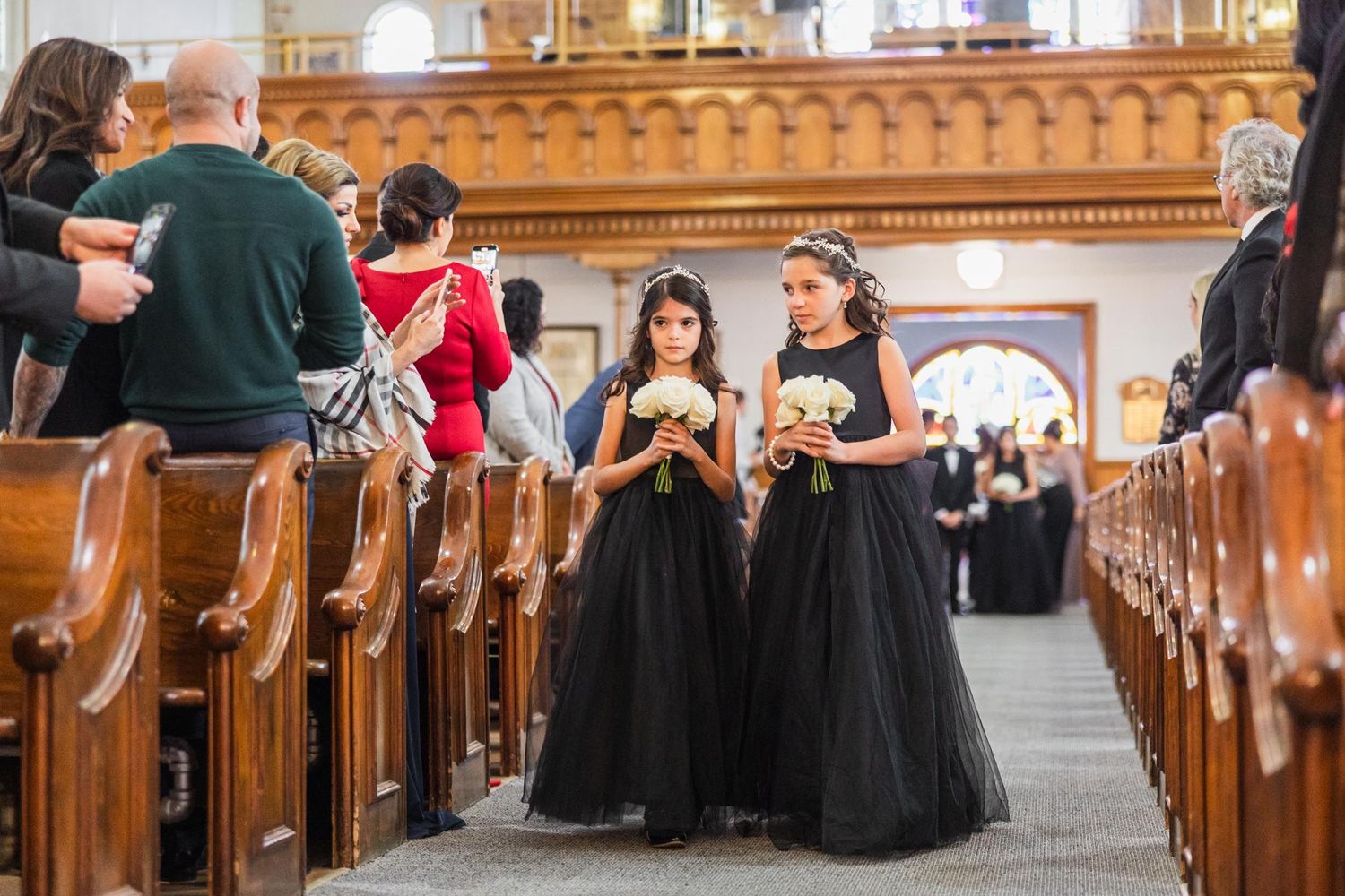 Two flower girls in black tulle dresses stand in church aisle during wedding ceremony.