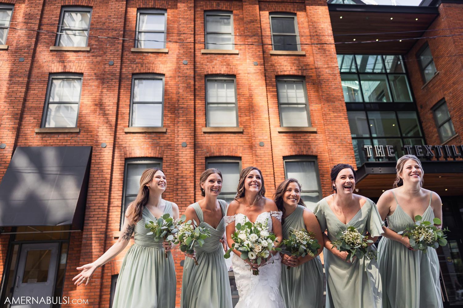 Bridesmaids in sage green dresses posing with bouquets against red brick wall.