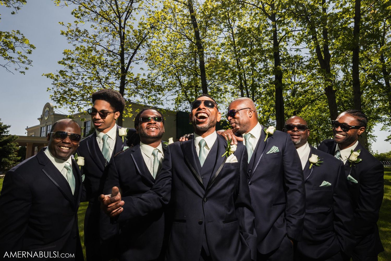 A wedding party of groomsmen in black suits and sunglasses pose outdoors in a fun group photo.