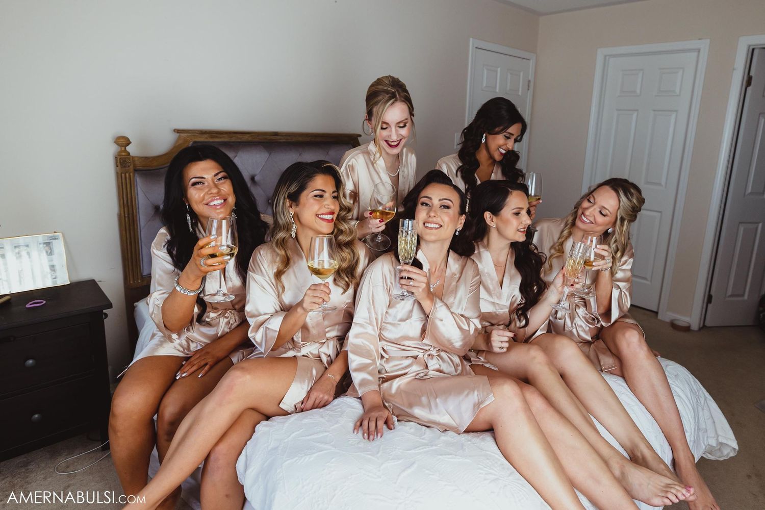 Group wearing matching silk robes sitting on bed celebrating with champagne.