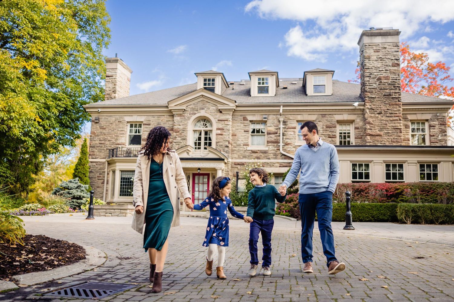 A family holding hands while walking in front of a grand stone mansion with autumn trees.