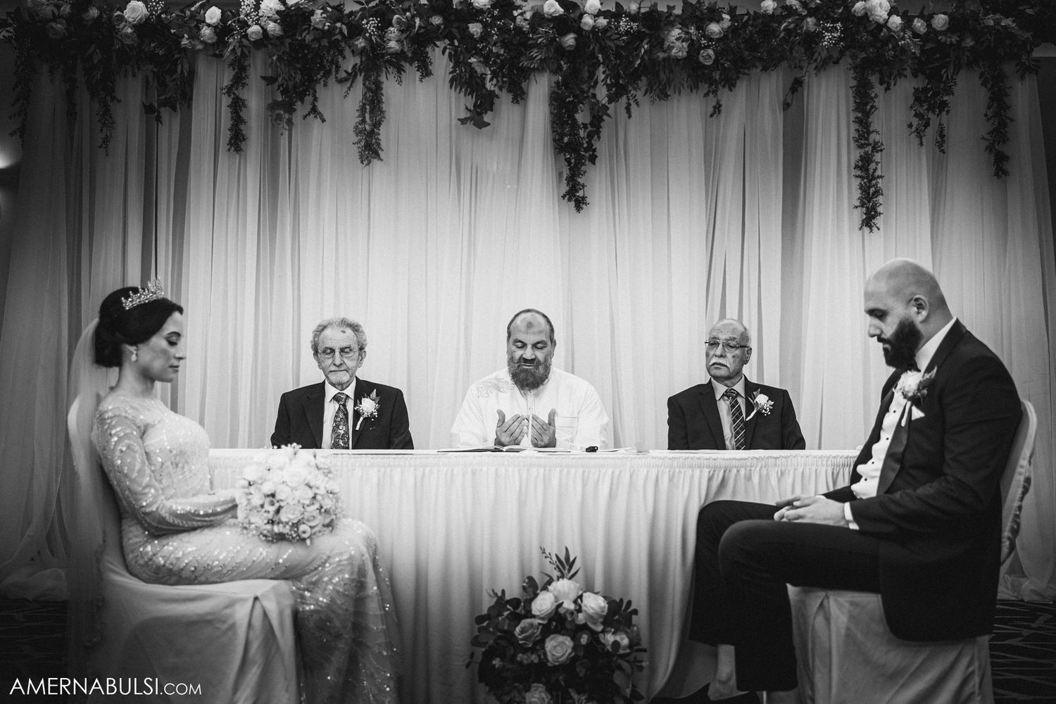 Black and white photo of a wedding ceremony with clergy and attendants at a decorated head table.